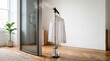 © Tina - A garment steamer is positioned next to a mirror in a bright room. A white shirt hangs on the steamer, with steam rising from it. A green plant is visible in the background.