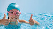 © Przemek - Happy young girl wearing a blue swimming cap and pink goggles giving a double thumbs up while playing in a sunny outdoor pool