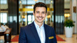 © Roman - a smiling man in a blue suit jacket and white shirt at a hotel lobby reception desk, slightly above eye level, with a welcoming and professional demeanor, showcasing a clean and modern look.
