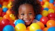 © Maryna - Curly haired Black child smiles brightly, buried in colorful ball pit. Kid plays happily amidst red yellow blue orange plastic spheres. Joyful toddler fun. Active indoor leisure.