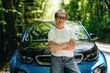 © standret - Middle-aged woman with short gray hair wearing glasses stands confidently with arms crossed in front of a blue electric car on a tree-lined road
