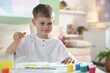 © New Africa - Little boy drawing picture with paints and brush at white desk indoors, space for text