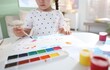 © New Africa - Little girl drawing picture with paints and brush at white desk indoors, closeup