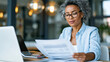 © Olha Havelia - Tax preparation concept, businesswoman in smart casual attire reviewing printed financial statements at a modern office desk, calculator and laptop open beside her, warm daylight t