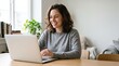 © Eve - Smiling young woman with wavy brown hair working on laptop at desk in cozy room. Happy female freelancer or student studying online, using wireless earphones and drinking coffee.