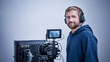 © ольга  - Man wears headphones and a blue hoodie. He stands beside a video camera with a monitor. The gray backdrop emphasizes the equipment. His smile shows approachability. Ideal for media production projects