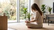 © Erika - Woman practicing mindfulness meditation and yoga poses sitting cross legged on rug in cozy living room with large windows overlooking green garden nature.