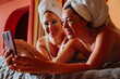 © oneinchpunch - Two happy women wearing under-eye patches and towels using smartphone in bed