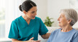 © SYATRI RAWU - Compassionate young nurse in teal scrubs talking to elderly woman at home, providing home healthcare and medical assistance to senior patient.