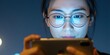 © Vadym - Asian woman wearing glasses uses smartphone in dark room. Blue light from screen reflects on glasses and face. She intently reads or texts on mobile device. Young adult focused on phone.