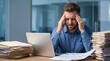 © ASyen - A stressed and overworked businessman holding his head in frustration while sitting at a desk piled high with documents