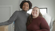 © Krakenimages.com - Man in grey turtleneck with arm around curvy woman in maroon top waving hand beside stacked moving boxes in a building interior; love together joy home.
