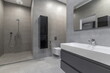 © Alhim - A bathroom features a large shower with a water fixture, a sink with a faucet, and a toilet. Gray tiles cover the walls and floor. The space has clean lines and a simple design