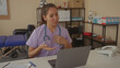 © Krakenimages.com - Young latina woman doctor with stethoscope speaking to laptop while touching chest in clinic building; telemedicine concern.