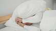 © Krakenimages.com - Woman lying in bed pulls pillow over head with one hand, face partially covered, white bedding and bedside plant visible in building; morning frustration.