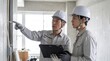 © k photo office - 建設現場で配線設備を指差し若手の施工管理士の人材育成を行う現場監督