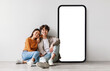 © Prostock-studio - Loving asian couple sitting near big cellphone with empty white screen, embracing, recommending newest mobile application or cool website over grey wall, mockup