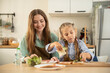 © sutthinon602 - Cute girl using both hands to build a sandwich while her smiling mother helps in the kitchen. Happy mother and daughter preparing healthy breakfast sandwiches together on a wooden cutting board.
