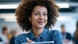 © Yassine - Smiling young woman with voluminous curly hair holds a digital tablet while working in a bright modern office environment today