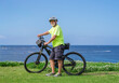 © luciano - Senior man with e-bike stopping on a coastal path overlooking the ocean, enjoying an active, healthy retirement lifestyle and freedom by the sea under a clear sky