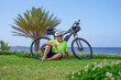 © luciano - Senior man cyclist relaxing on grass near electric bicycle, enjoying retirement and healthy active lifestyle by the ocean with tropical palm trees in background