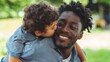 © Olya Pilipchik  - African american father smiling while carrying young son piggyback, receiving a loving kiss on the cheek, celebrating family bonds and parenthood outdoors