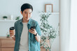 © Monster Ztudio - Happy Asian man drinking hot coffee in the morning using the phone while standing in the living room at home. Handsome young man walking around with a cup of hot drink, looks at a smartphone