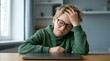 © Yassine - Stressed young boy wearing black glasses sits at a wooden table with a closed laptop looking tired and frustrated today