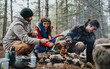 © qunica.com - Three friends roast sausages over a small campfire while sitting at a forest campsite. They laugh and cook together beside a tent during a chilly outdoor camping trip.