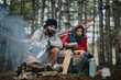 © qunica.com - A couple sits by a smoky campfire in a pine forest cooking and preparing food. They relax near a colorful hammock while arranging fruit and snacks on a rock.