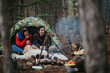 © qunica.com - A young couple sits inside a camouflage tent holding mugs while their dog lies nearby by a small campfire. The pair enjoy a cozy camping moment together in a quiet pine forest setting.