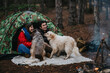 © qunica.com - A young couple sits inside a camouflage tent with two dogs at a forest campsite. They laugh and interact with their pets beside a campfire and backpacks.