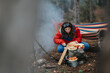 © qunica.com - A woman in a red jacket prepares bread and fruit on a rock at a campsite. She arranges food next to cookware and a colorful hammock while a dog watches.