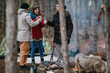 © qunica.com - Three friends stand by a campfire sharing hot drinks during a forest camping trip. The group enjoys a relaxed outdoor moment beside their tent and gear.