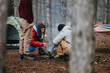 © qunica.com - Three friends make a small campfire while camping in a pine forest. A group of campers crouch around the fire next to a tent and hammock.