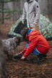 © qunica.com - Two friends are camping in a mountain forest with a dog beside a camouflage tent. One person crouches to interact with the dog while the other stands near the tent.
