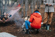 © qunica.com - Three people are camping in a pine forest gathered around a small campfire. One person squats tending the fire while another relaxes in a hammock and a third stands nearby.