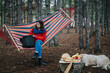 © qunica.com - A young woman sits in a colorful striped hammock in a pine forest holding a backpack. A relaxed outdoor camping scene shows her resting while a dog lies nearby by picnic food.