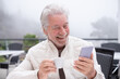 © luciano - Senior white-haired man laughing and smiling while looking at his smartphone, enjoying a cup of coffee at an outdoor cafe on a foggy day, embracing technology and leisure