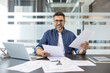 © Liubomir - Experienced businessman smiling at camera, sitting at office desk with laptop and documents, analyzing reports, and managing business tasks with confidence and expertise