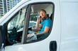 © Videophilia - A man sits inside a delivery van parked on a city street. He uses a phone to talk while wearing a headset. Boxes are placed in the van. It is bright and sunny outside.
