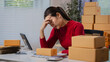 © NanSan - Tired small business owner in a red sweater sits at a desk with shipping boxes, looking stressed while working on online orders in a home office.