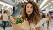 © pattozher - Shocked Shopper: A woman with a distressed expression clutches a paper bag filled with groceries, looking at a receipt with disbelief, while navigating a crowded shopping mall.