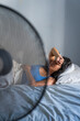 © davide bonaldo - young adult woman in bed during a hot summer heat wave with fan, cooling off fighting heat at home during a heatwave and high temperature