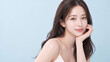 © YT - 淡いブルー背景と広い余白のあるスタジオで頬に手を添えてやさしく微笑むロングヘアの若いアジア人女性、透明感のある美容・スキンケア広告向けビューティーポートレート
