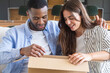 © simona - Happy mixed-race couple sitting on a couch excitedly unboxing a cardboard package together at home, sharing a surprised smile and warm moment of connection and anticipation.