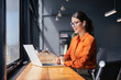 © Stock 4 You - Smiling young latin or indian businesswoman wearing glasses working on laptop computer at wooden desk by window. Professional woman typing, listening music in earphones in modern coworking office.