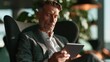 © CYBERPINK - Middle-aged man sitting in a chair and using a tablet. he is wearing a grey blazer and a white shirt. he has a beard and is looking down at the tablet with a serious expression on his face.