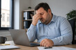 © Anastasiia - A middle-aged man in a blue shirt is sitting at a desk, looking stressed and holding his head while working on a laptop in an office setting.