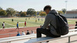 © Nikolay - A young African American man sits on bleachers, observing a lively game on the athletic field. This depicts youth education and campus life, showcasing a moment of thoughtful contemplatio